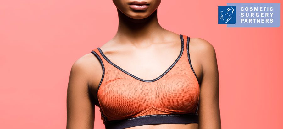Woman wearing sports bra showing natural breast shape on pink background – Cosmetic Surgery Partners logo in corner.