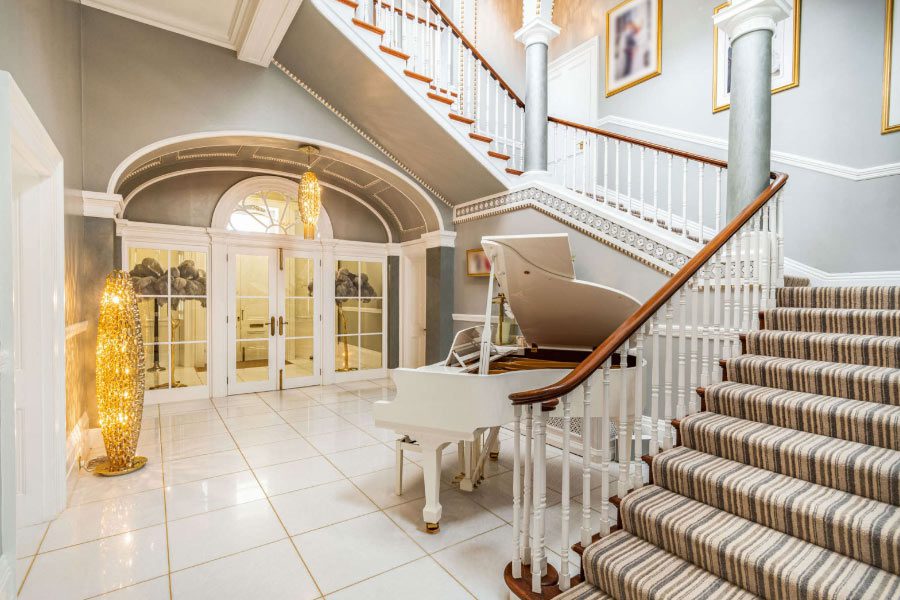 Entrance Hall: The opulent marble entrance hall at Cosmetic Surgery Partners' Hemingford Park suite, featuring a grand piano and sweeping staircase.
