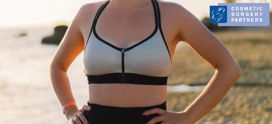 Woman standing on beach wearing sports bra after breast surgery recovery – Cosmetic Surgery Partners logo in corner